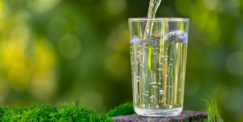 Bicchiere d'acqua pura versata all'aperto su sfondo verde naturale, simbolo di sostenibilità e tecnologie per l'estrazione dell'acqua dall'aria.