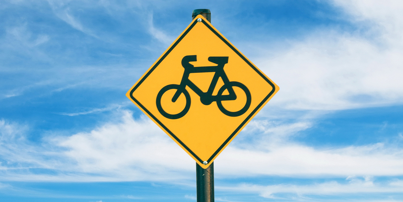 Cartello giallo che segnala un attraversamento ciclabile, con cielo azzurro sullo sfondo.